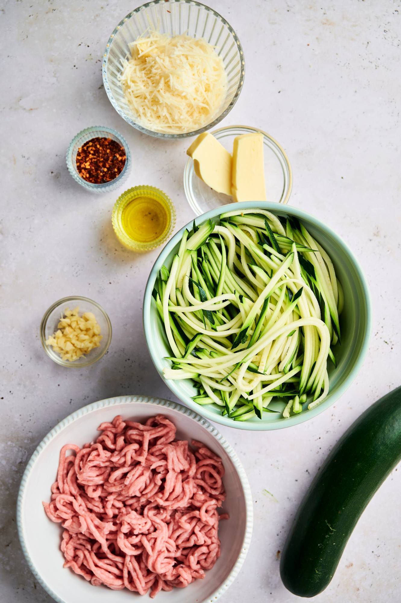 Ingredients for zucchini pasta including ground sausage, zuchini noodles, garlic, olive oil, butter, and Parmesan cheese.