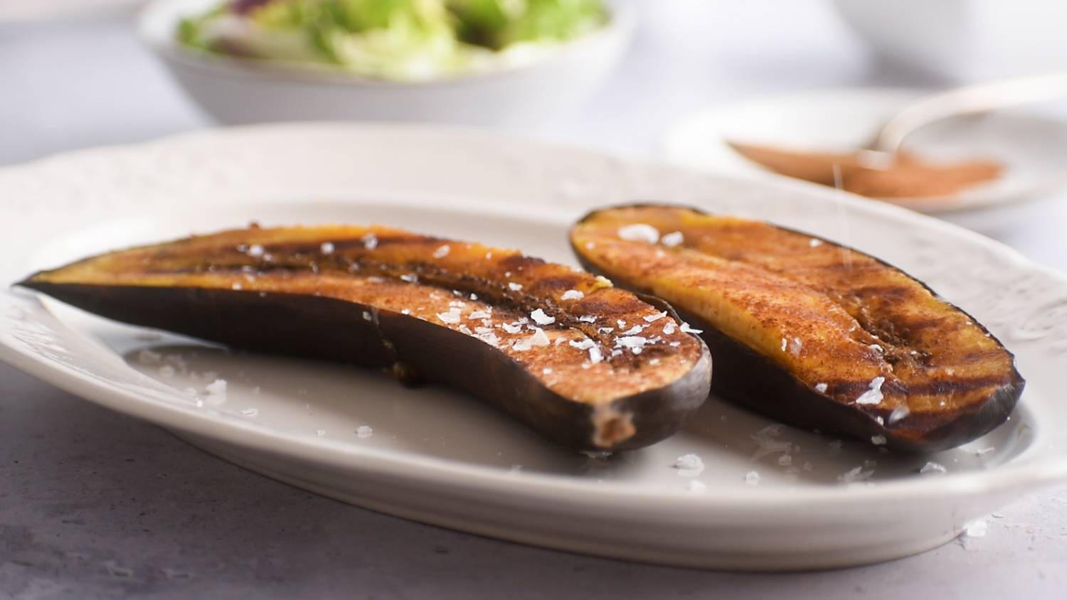 Two roasted plantains seasoned with salt, placed on a decorative plate, with a fresh salad in the background.