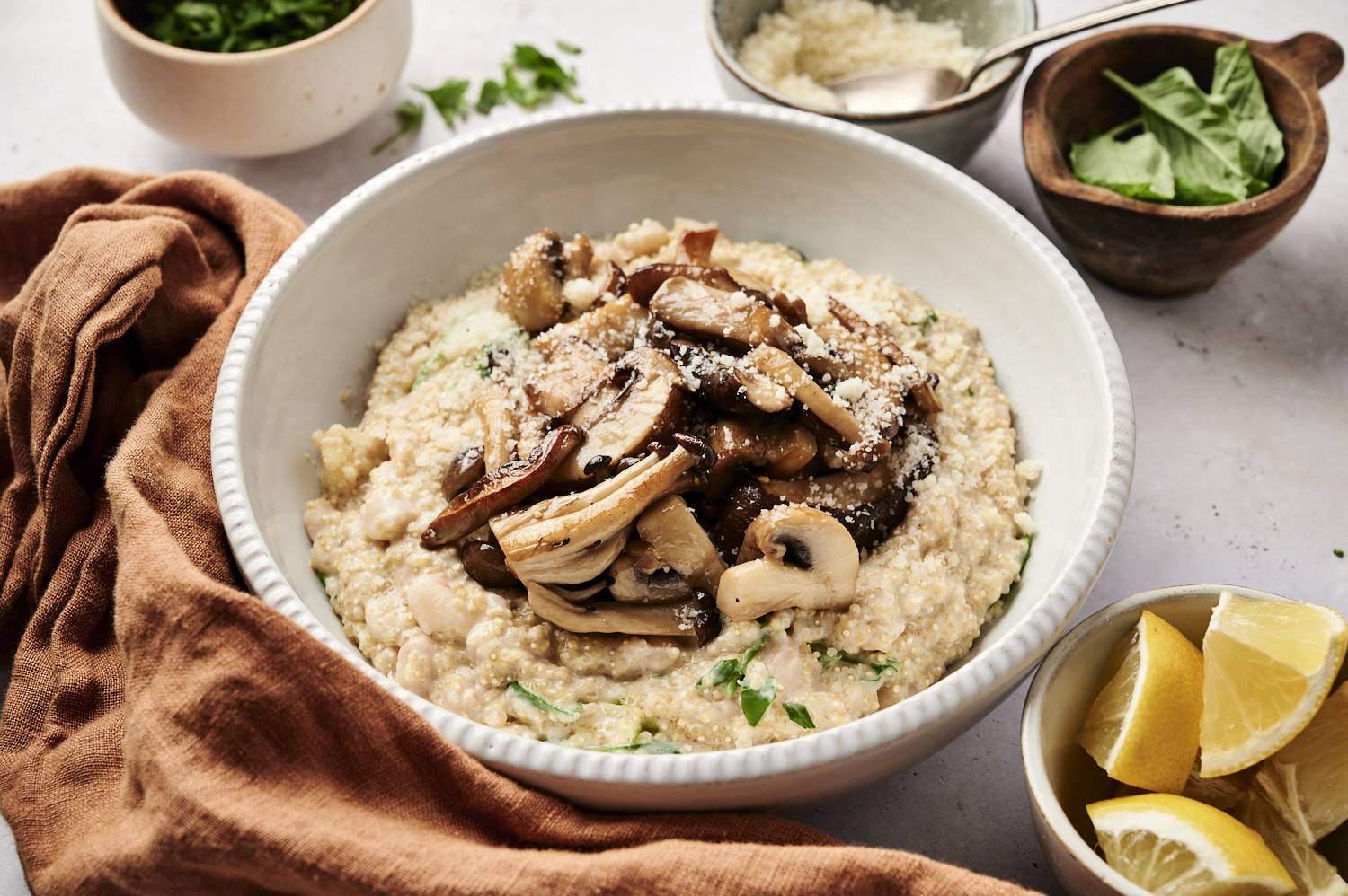 A creamy quinoa dish topped with sautéed mushrooms and Parmesan, served in a white bowl with fresh basil and lemon wedges on the side.