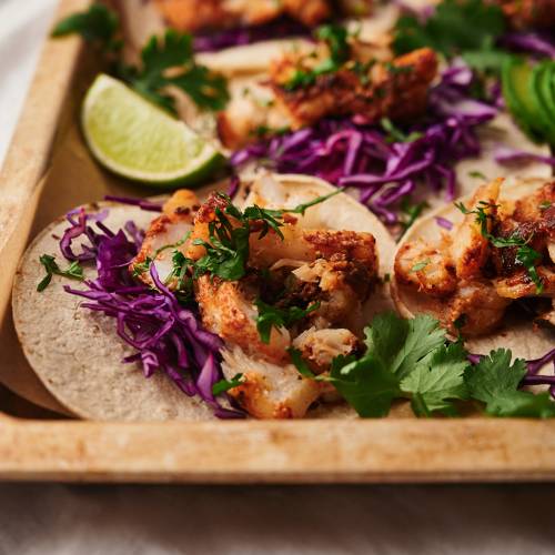 Fish tacos with flaky seasoned fish served in a corn tortilla with cilantro, shredded red cabbage, and limes.