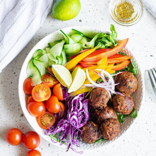 Lentil meatballs served in a bowl with cabbage, peppers, tomatoes, cucumber, red onion, and limes.