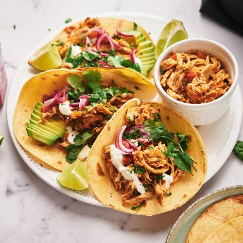3 Shredded chicken tacos with avocado, pickled onions, cilantro, and limes served on a round plate.