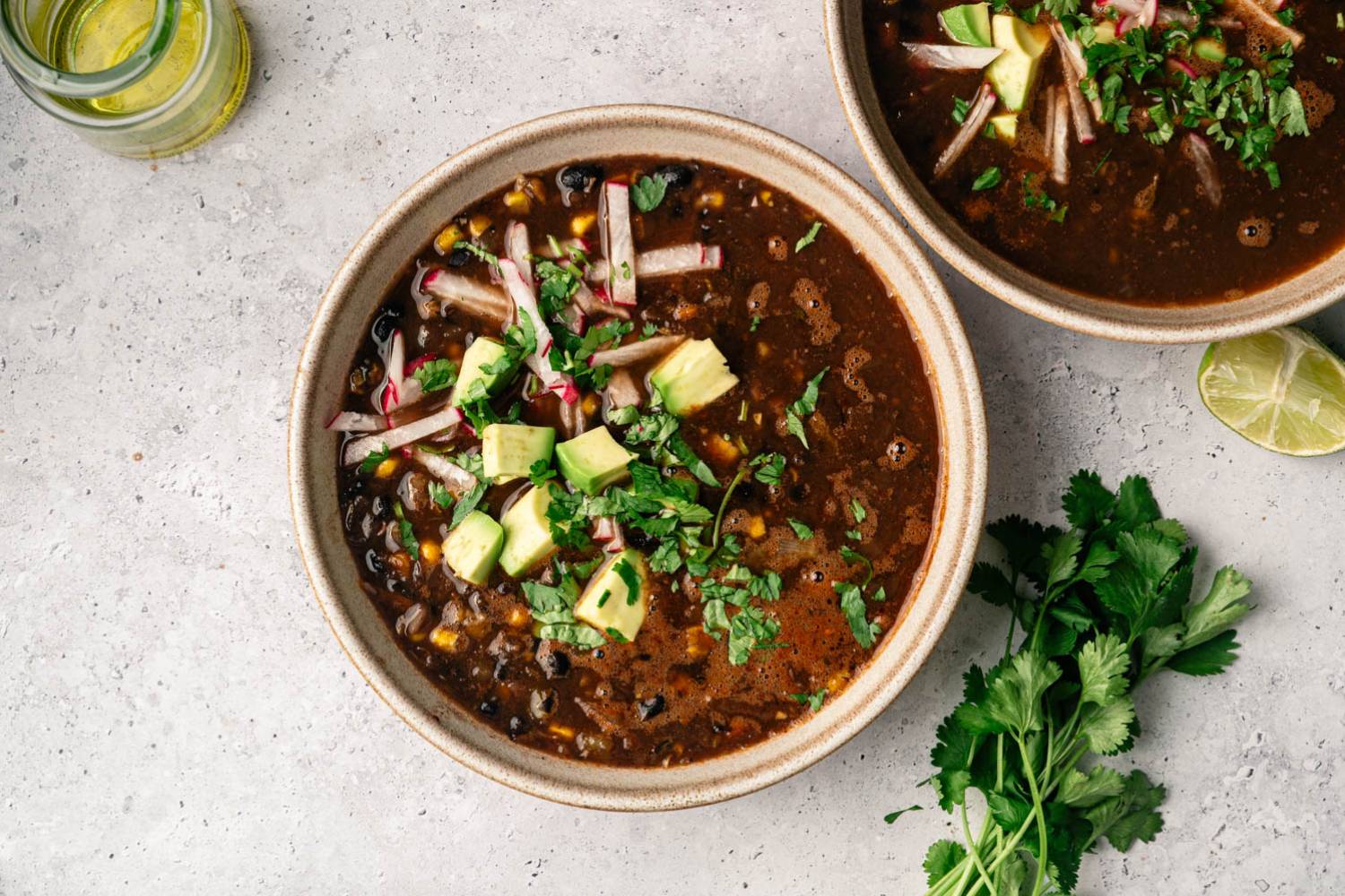 A bowl of black bean soup topped with avocado, cilantro, and radish strips on a textured gray surface. A jar of oil and fresh lime and cilantro are nearby.