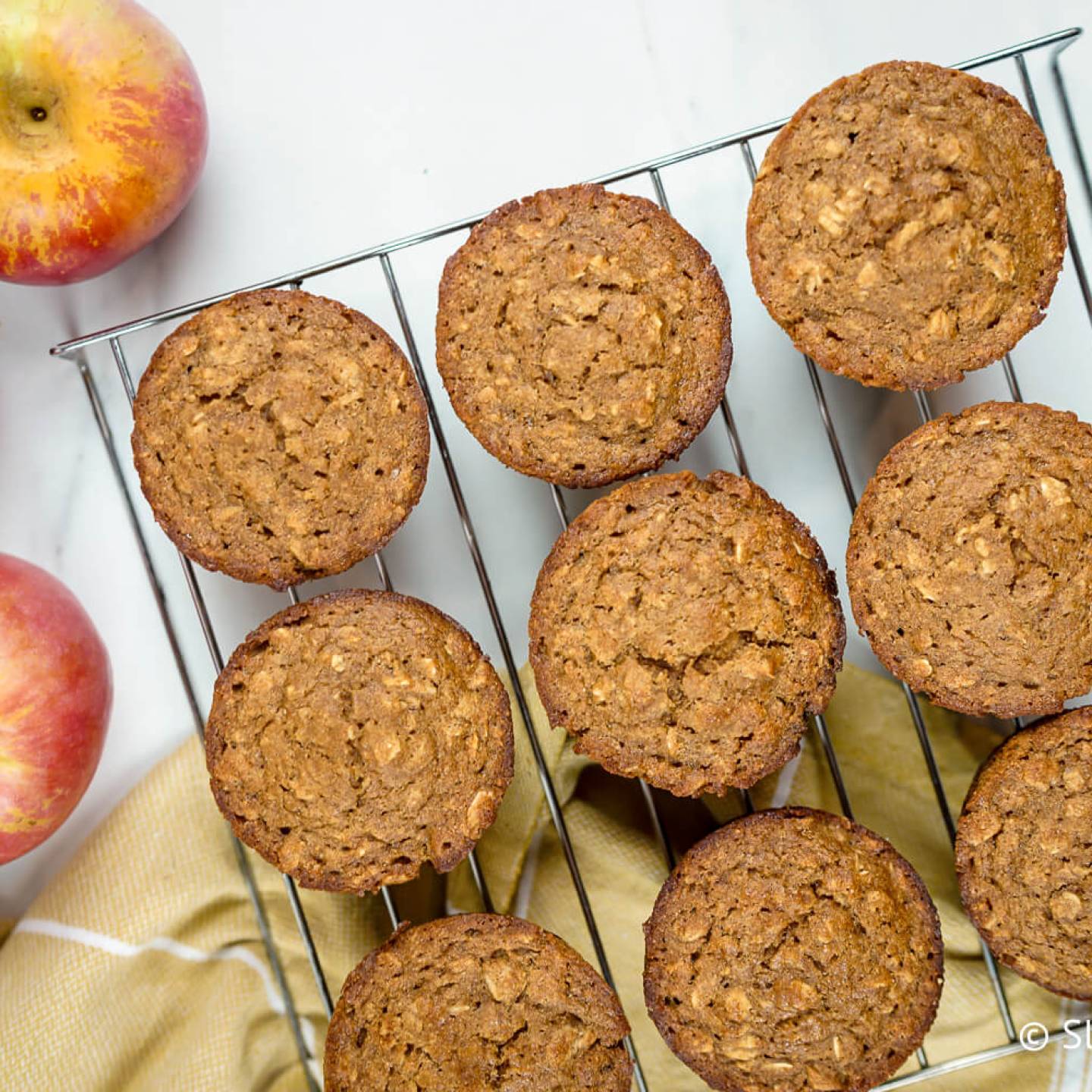 Applesauce oatmeal muffins with oatmeal flecks on a baking rack with a yellow napkin and apples.