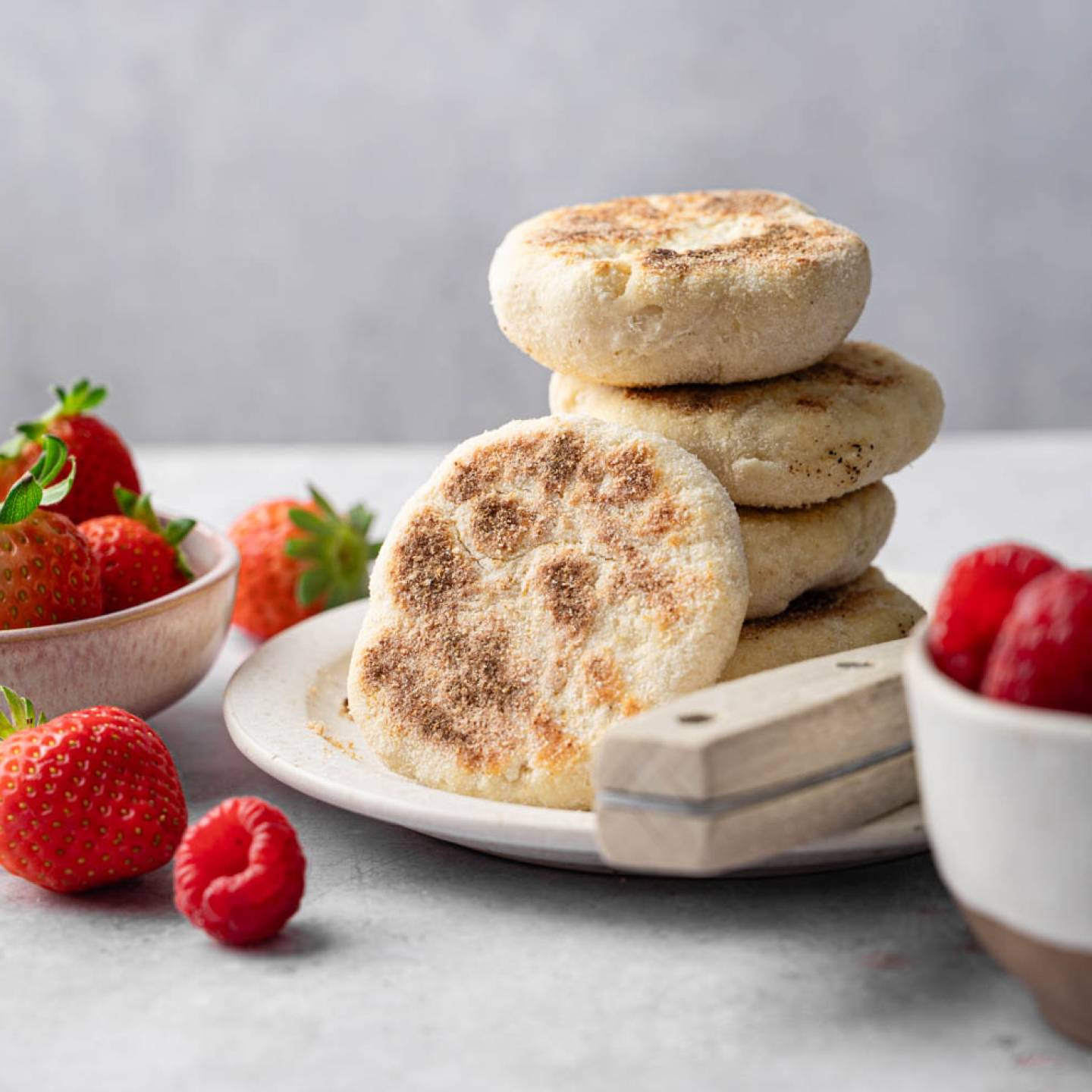 A plate with stacked English muffins and a cutting board on a gray surface. Nearby bowls hold fresh strawberries and raspberries, creating a fresh and inviting scene.
