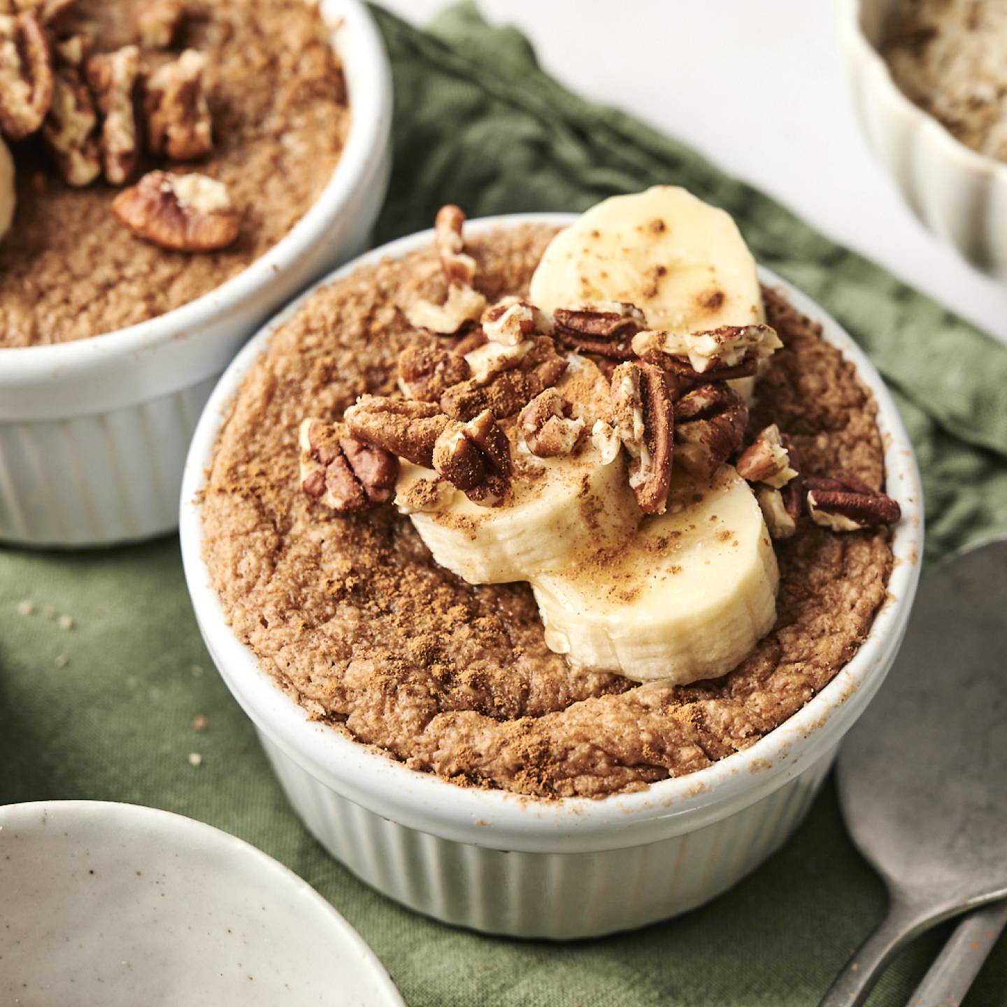 Two ramekins of baked protein oats topped with sliced bananas, chopped pecans, and a sprinkle of cinnamon, placed on a green napkin with a bowl of oats and spoons in the background.