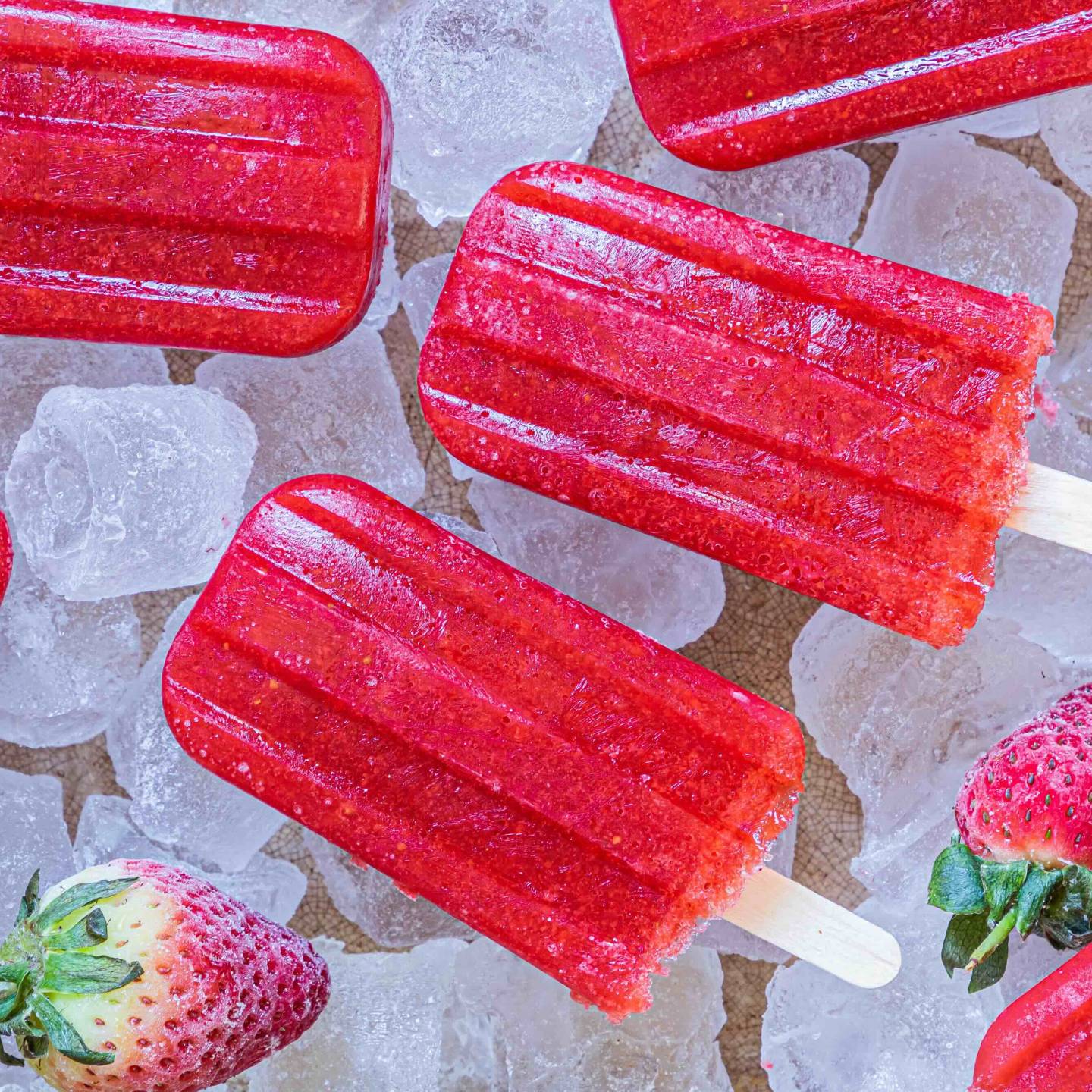Bright red strawberry popsicles on ice, accompanied by fresh strawberries, create a refreshing summer treat scene.