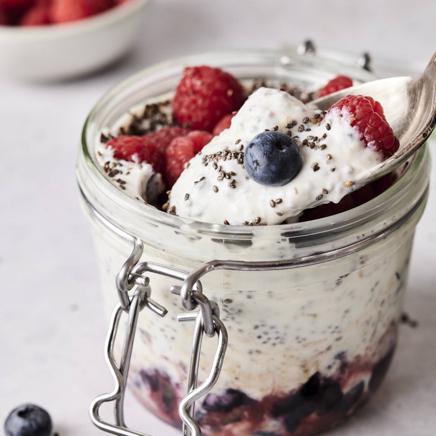 Spoon scooping creamy yogurt chia seed pudding topped with raspberries, blueberries, and chia seeds from a glass jar layered with fruit at the bottom.