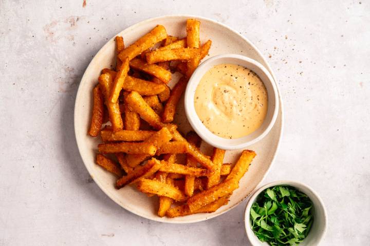 Plate of crispy golden fries with a creamy dipping sauce on the side. A small bowl of fresh green parsley is nearby on a light, textured surface.