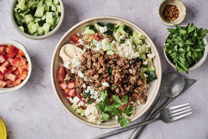 Bowl of hummus topped with seasoned ground meat, cucumber, tomatoes, feta, and fresh parsley, surrounded by ingredient bowls and utensils.