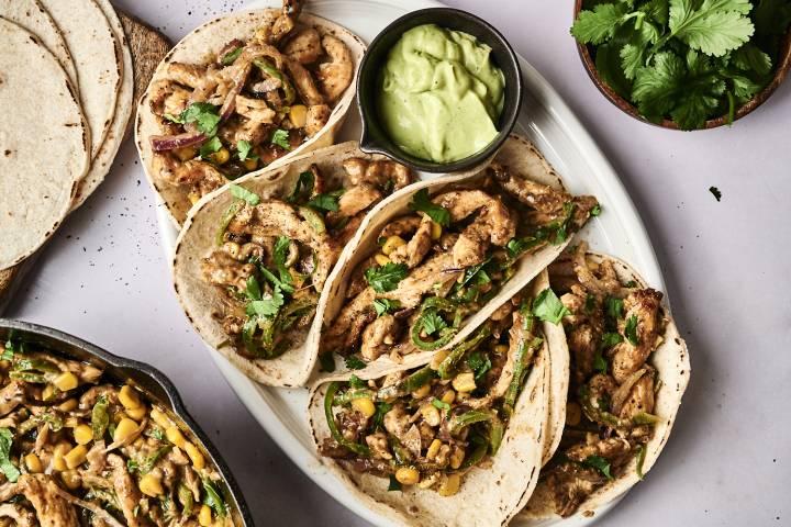A platter of chicken tacos topped with cilantro and corn, accompanied by a bowl of green sauce, on a light background.