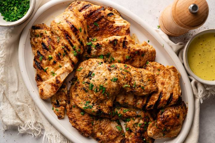 Grilled chicken breasts garnished with chopped parsley on a white plate. A small bowl of green herbs and a bowl of yellow sauce are nearby.