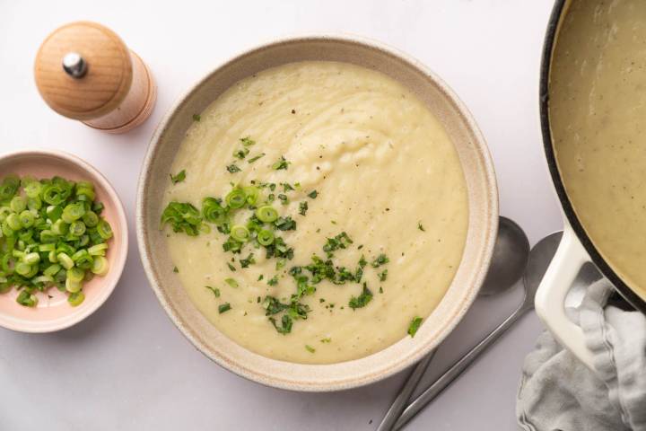 Creamy potato soup in a bowl garnished with fresh herbs and green onions. Nearby are a pink dish with green onions, a pepper mill, and a ladle beside a pot.