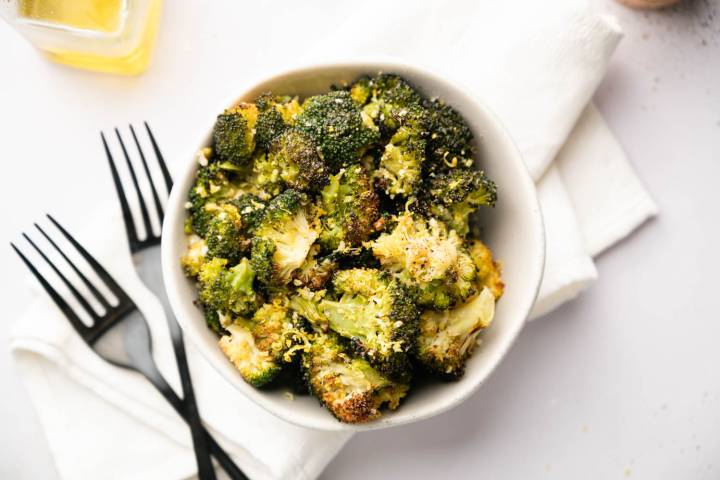 Bowl of roasted broccoli topped with grated cheese, set on white napkin. Two black forks lie beside, with a glass of yellow liquid nearby.