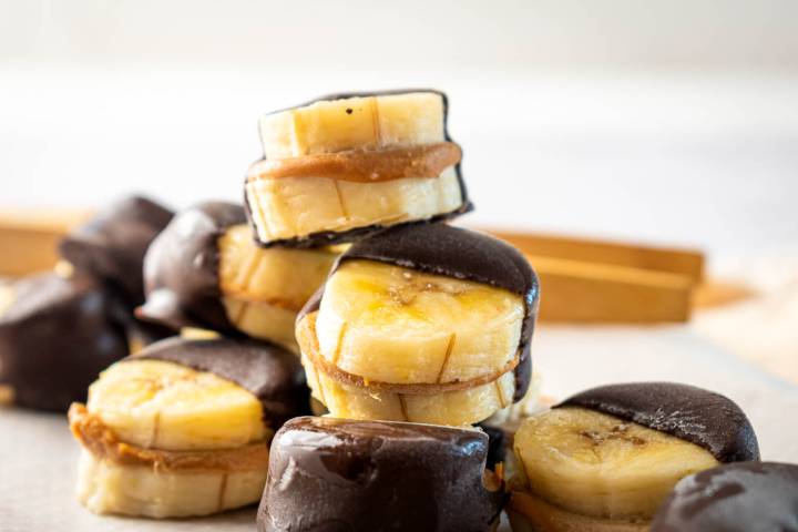 Stacked banana slices filled with peanut butter, dipped in dark chocolate, arranged artfully on a light background.