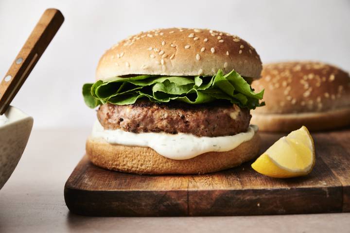Greek turkey burger on a wooden board with crisp lettuce and creamy tzatziki sauce inside a sesame seed bun, garnished with a lemon wedge on the side.
