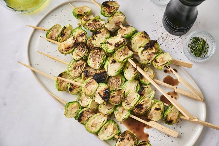 Grilled Brussels sprouts skewered and arranged on a white platter, drizzled with balsamic glaze and sprinkled with cracked black pepper, with rosemary and olive oil in the background.