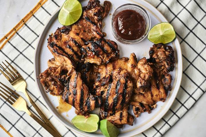 Grilled chicken thighs garnished with lime wedges served on a plate with a side of barbecue sauce. Elegant utensils nearby.
