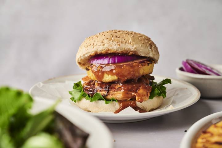 A delicious sesame seed burger with grilled chicken, lettuce, a slice of pineapple, red onion, and a savory sauce on a white plate.