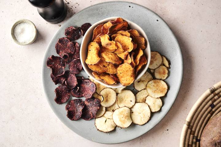 A platter of assorted vegetable chips in three colors: purple, golden, and white, served alongside a small bowl of salt.