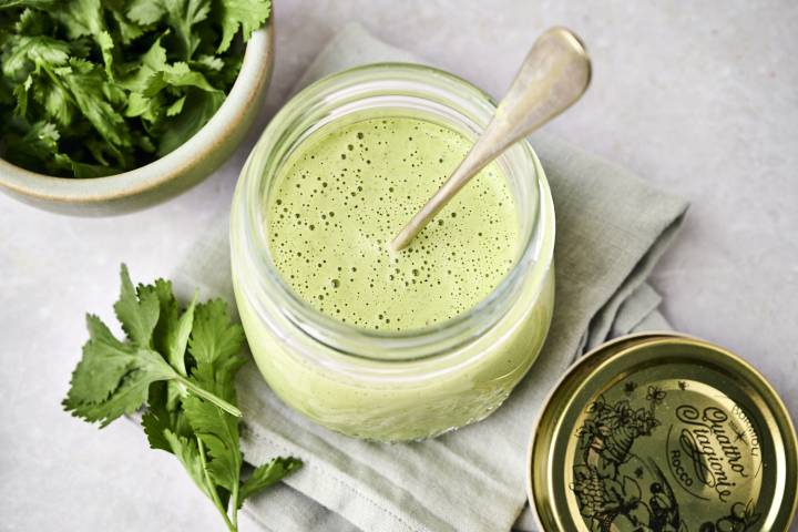 A jar of green smoothie with bubbles and a spoon, beside a bowl of fresh cilantro on a cloth.