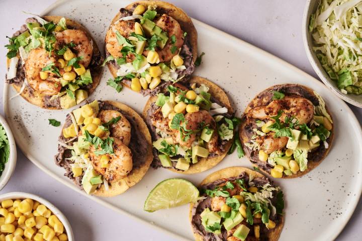 A plate of six shrimp tostadas with black beans, shredded cabbage, corn, avocado, and cilantro.