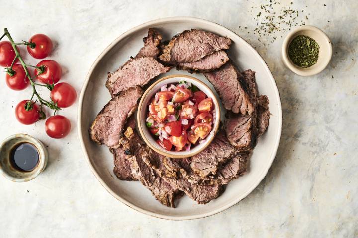 Platter of sliced beef with a bowl of fresh tomato salsa, surrounded by cherry tomatoes and spices on a light background.