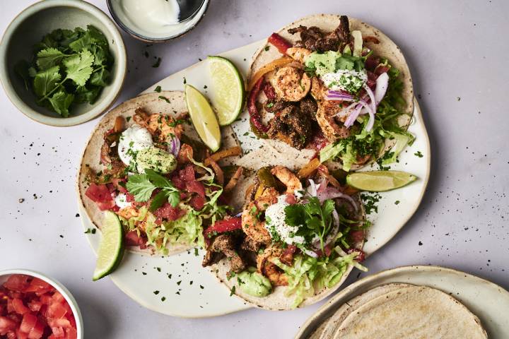Three vibrant tacos topped with shrimp, beef, lettuce, tomatoes, onions, and cilantro, served with lime wedges and dipping sauce.
