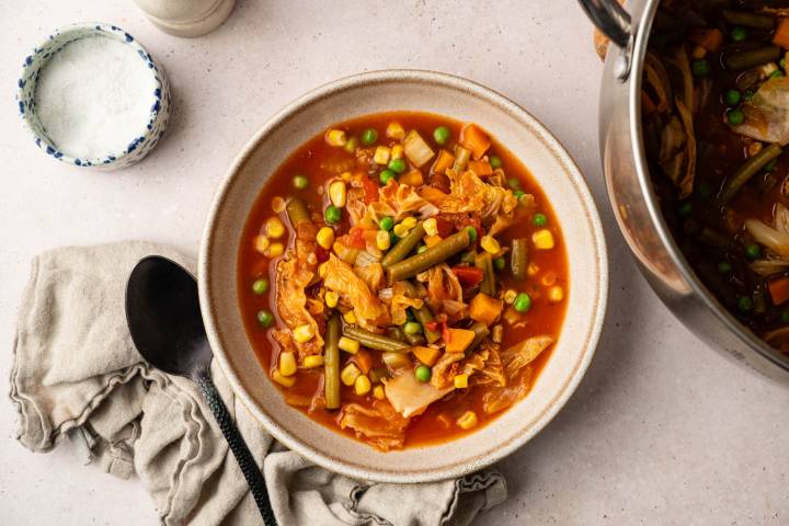 Bowl of vibrant vegetable soup with corn, beans, carrots, and cabbage in tomato broth. Nearby, a black spoon on a beige napkin, with a salt container.