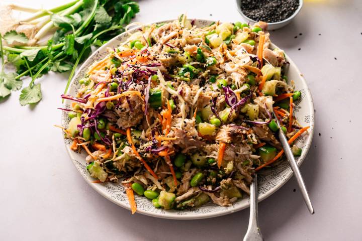 A vibrant Asian-inspired salad with shredded chicken, edamame, cucumber, carrots, and red cabbage on a plate, garnished with black sesame seeds.