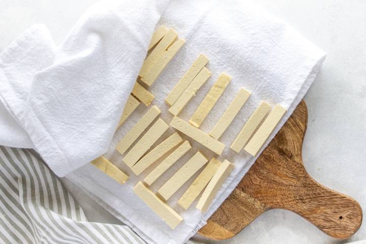 Tofu cut into strips on a cutting board with a kitchen towel.