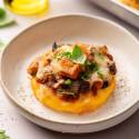 Polenta cake topped with tomato sauce, melted cheese, mushrooms, and herbs on a white plate. Fresh basil leaves and herbs surround the dish.