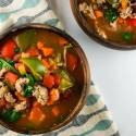 Sausage soup with peppers and spinach in a wooden bowl with carrots, celery, and tomato broth.