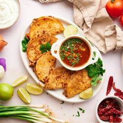 Chicken birria tacos with corn tortillas filled with melted cheese and served with consommé, cilantro, onion, and lime juice.
