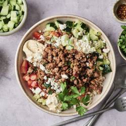 Bowl of hummus topped with seasoned ground meat, cucumber, tomatoes, feta, and fresh parsley, surrounded by ingredient bowls and utensils.