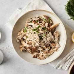 A bowl of shredded Greek chicken, garnished with fresh herbs. Surrounded by lemon, dill, cherry tomatoes, and salt, creating a fresh Mediterranean feel.