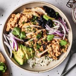 A bowl filled with grilled chicken, rice, black beans, avocado, red onion, and creamy sauce, garnished with fresh cilantro.