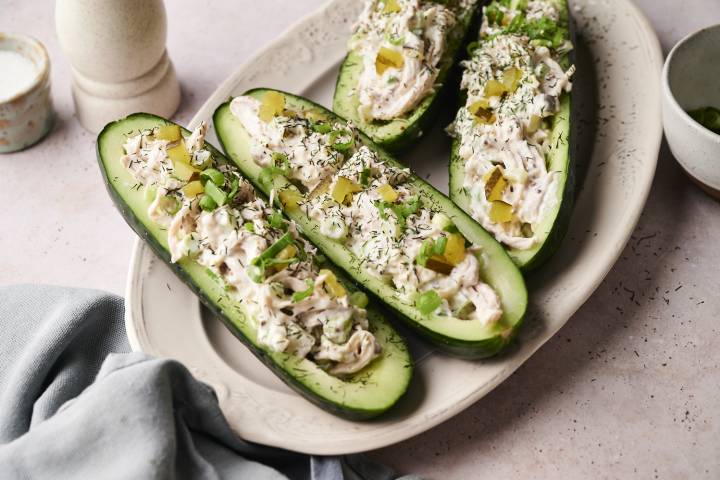 Four cucumber “boats” filled with creamy shredded chicken salad, garnished with chopped pickles, sliced green onions, and a sprinkle of dried dill, arranged on a white oval plate with a light blue cloth napkin nearby.