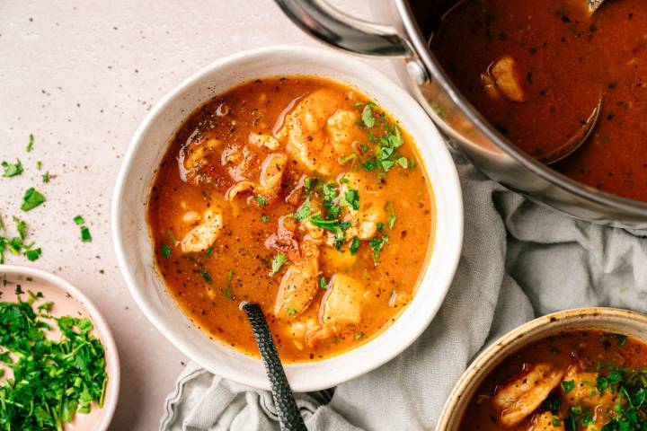 A bowl of hearty tomato soup with shrimp and herbs. The soup is a rich, warm reddish color, garnished with chopped parsley, next to a pot. Cozy and appetizing.