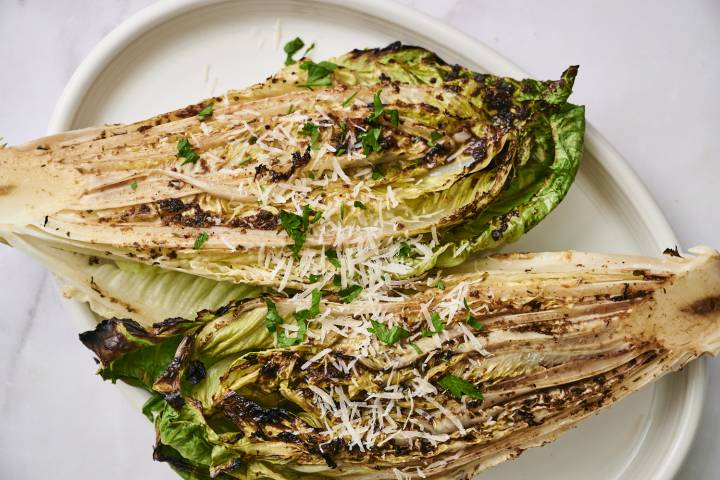 Grilled romaine lettuce halves topped with grated Parmesan, herbs, and black pepper, served with a lemon slice on a white plate.