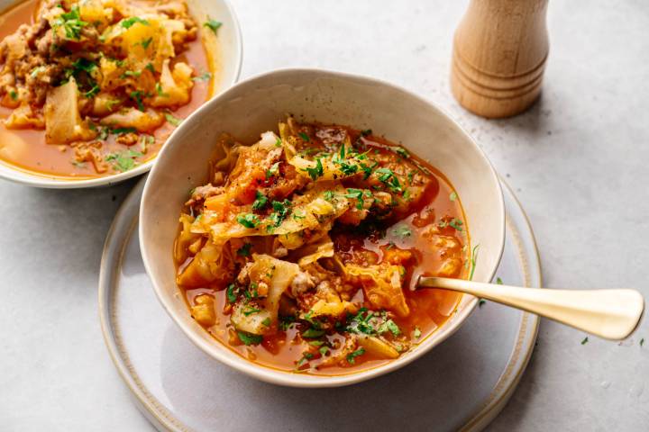A bowl of hearty cabbage soup with ground meat, garnished with fresh parsley, sits on a light gray surface. A second bowl and pepper mill in the background.