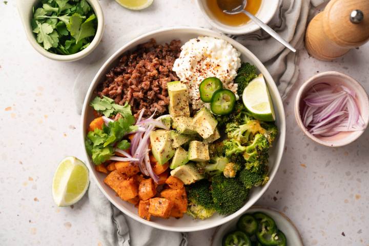 A vibrant bowl of mixed ingredients, including diced avocado, jalapeños, ground beef, broccoli, cottage cheese, red onion, and sweet potato. Garnished with cilantro and lime wedges on a speckled surface, conveying a fresh and hearty meal.