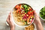 Woman hands holding a bowl with creamy hummus, quinoa, chickpeas, cucumbers, arugula, and red onions.