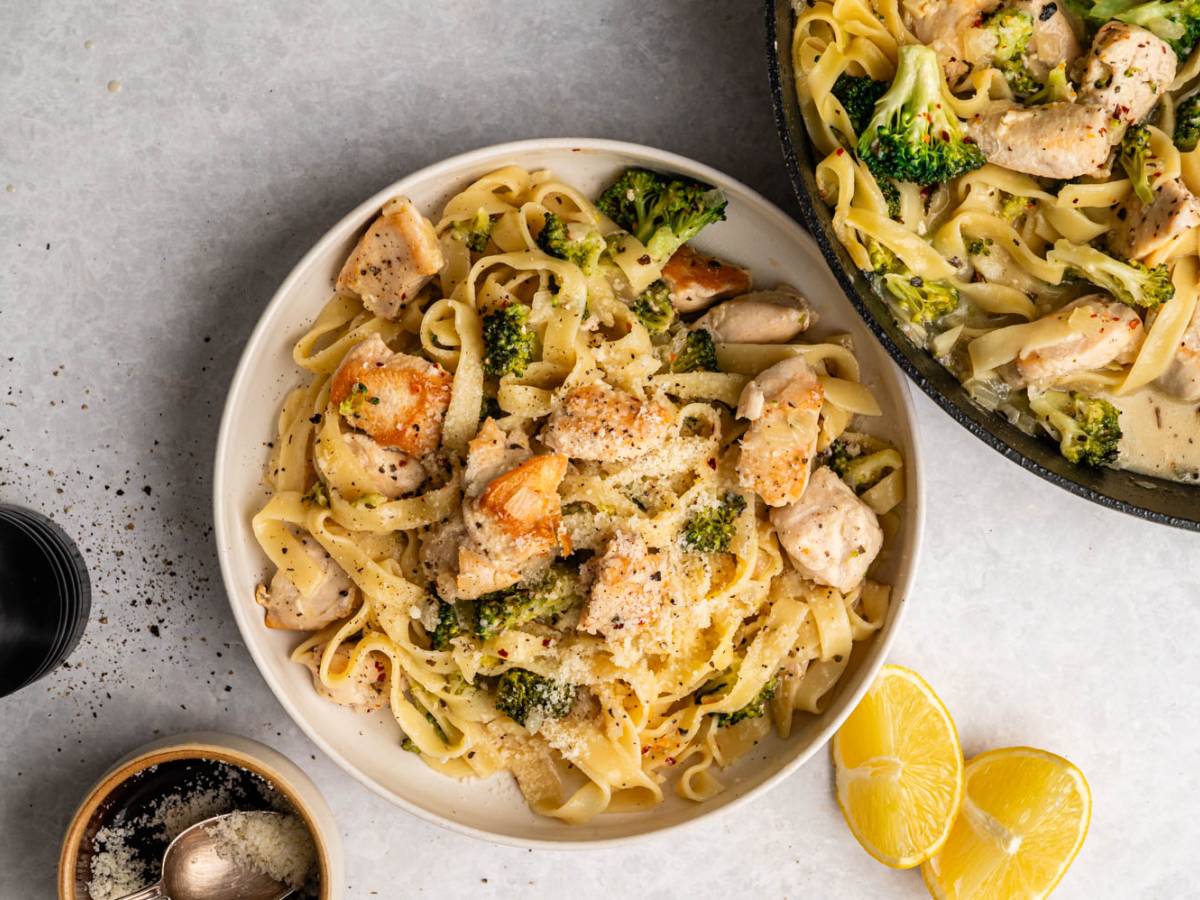 Plate of creamy chicken and broccoli pasta topped with parmesan, next to a similar skillet. Lemon wedges and pepper grinder add fresh vibes.