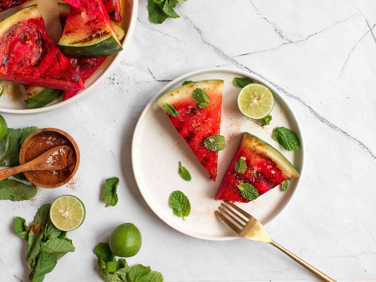 Slices of grilled watermelon on a white plate garnished with fresh basil, cut limes, and chili powder on the side.